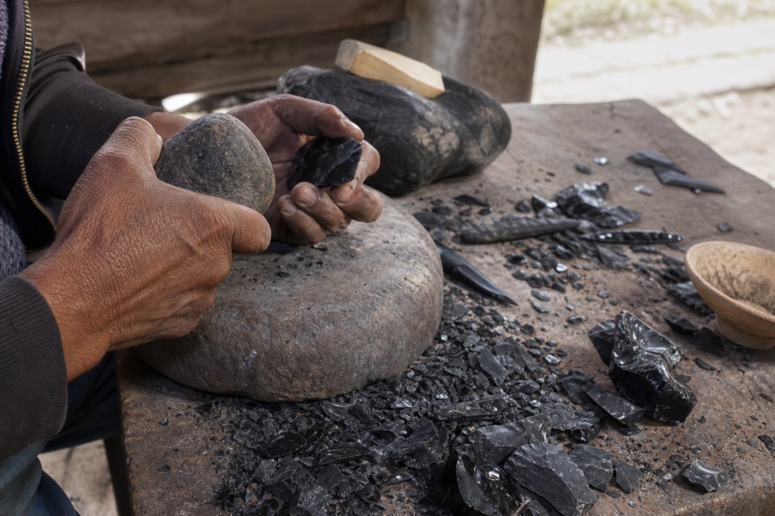 Hands demonstrating obsidian knapping technique over a workbench